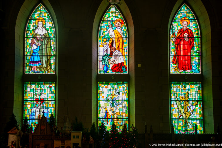 Three tall stained-glass windows with sacred figures and symbolic lower panels glowing above Christmas decorations.