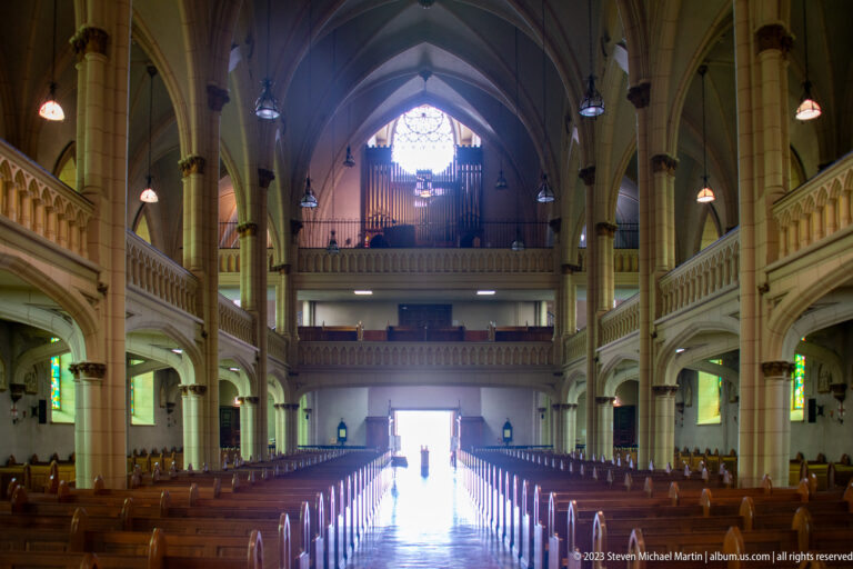 Rearward view down a neo-Gothic church nave toward an organ loft, stained-glass window, side galleries, and bright doorway.