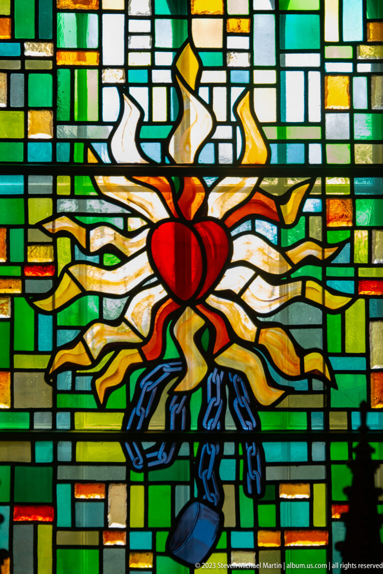 Stained-glass panel with a red Sacred Heart amid white and gold rays, blue ornament, and green geometric glass.