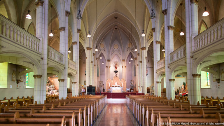 Long neo-Gothic nave viewed toward the illuminated altar, with pointed vaults, columns, side galleries, and wooden pews.
