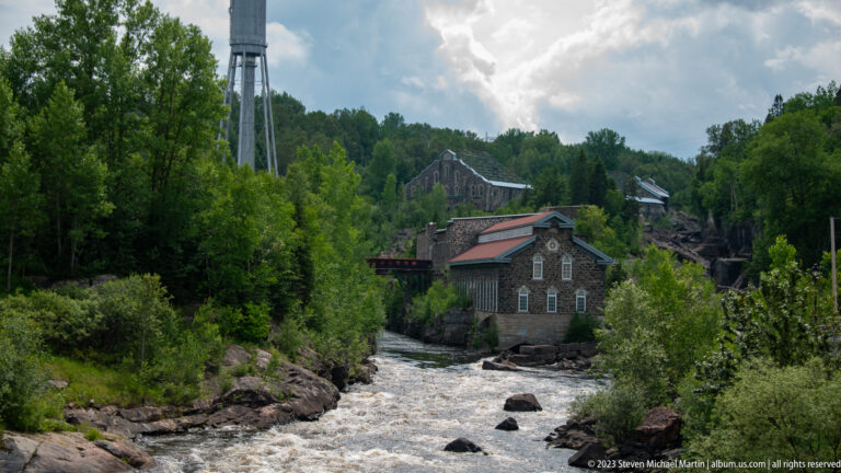 Stone industrial buildings with red roofs beside a fast rocky river, backed by dense green trees and a tall modern structure.