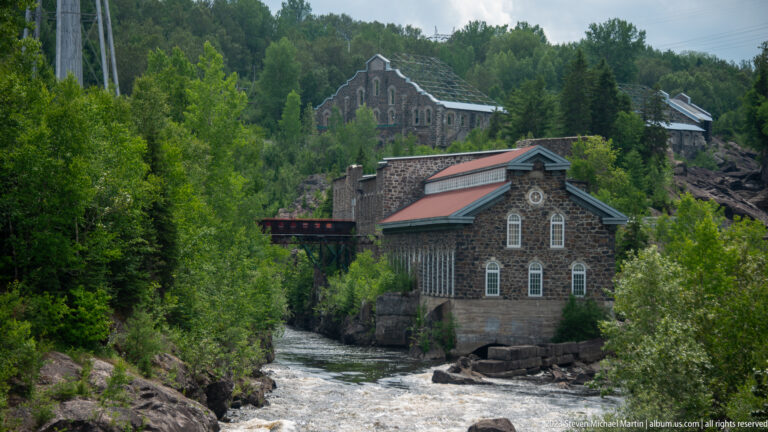 Stone industrial buildings with arched windows and red roofs beside a rushing river and wooded hillside.