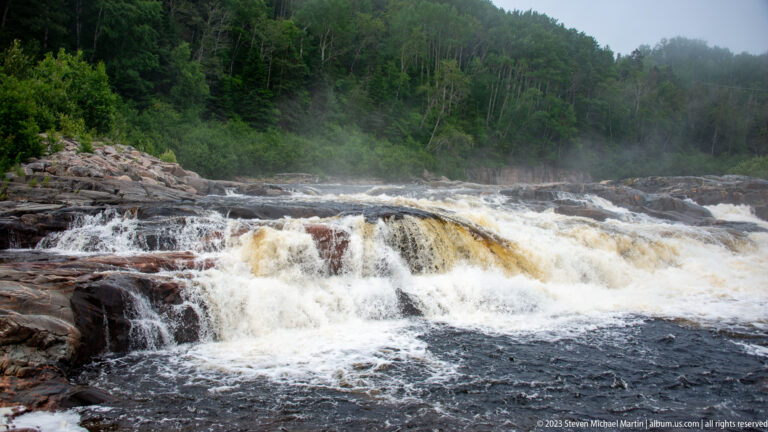 Broad amber-and-white cascade over dark rock shelves with mist and dense green forest behind.