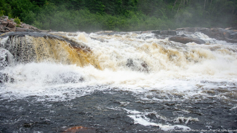 Close view of amber-tinted river water breaking into white foam over a broad dark bedrock ledge.