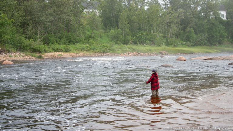 Person in a red plaid jacket standing knee-deep in a shallow river near boulders and a wooded bank.