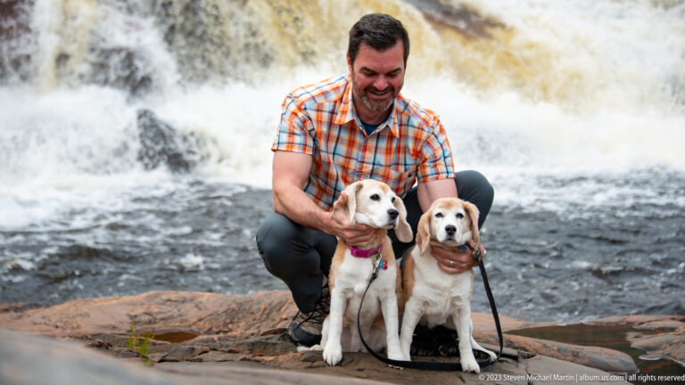 Smiling man crouching with two beagles on a rock ledge in front of foaming river rapids.