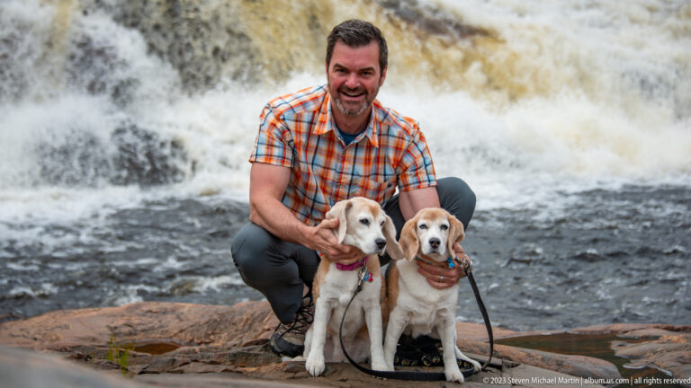 Man crouching behind two seated beagles, all facing the camera with foaming rapids behind them.