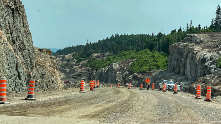 Gravel highway through a rock cut with orange construction barrels, workers, vehicles, and conifer forest.