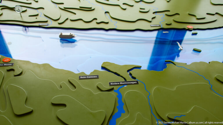 Raised relief map with blue water, green land, small boats, and labels including Baie-Comeau and Rivière Manicouagan.