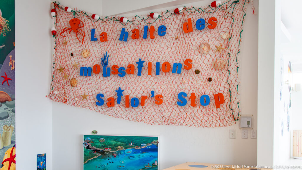 Orange fishing net with large blue and red exhibit lettering above a small underwater illustration in a white museum gallery.