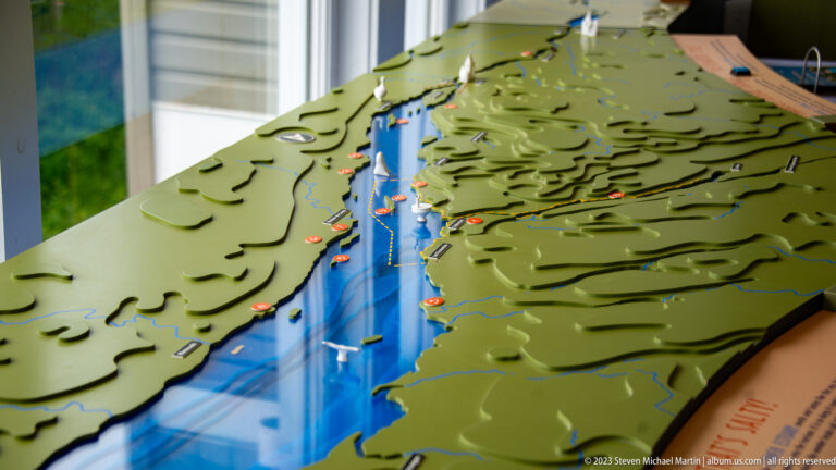 Raised green topographic model flanking a long blue estuary channel with small labeled markers and settlements.