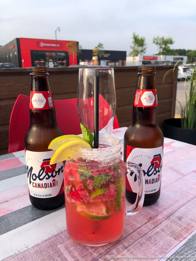 Two Molson Canadian beer bottles beside a red cocktail with lemon and herbs on a patio table.