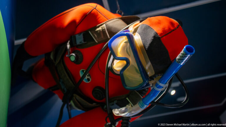 Red diving or flotation gear with a clear mask, blue snorkel, straps, hoses, and fittings in a dark exhibit.