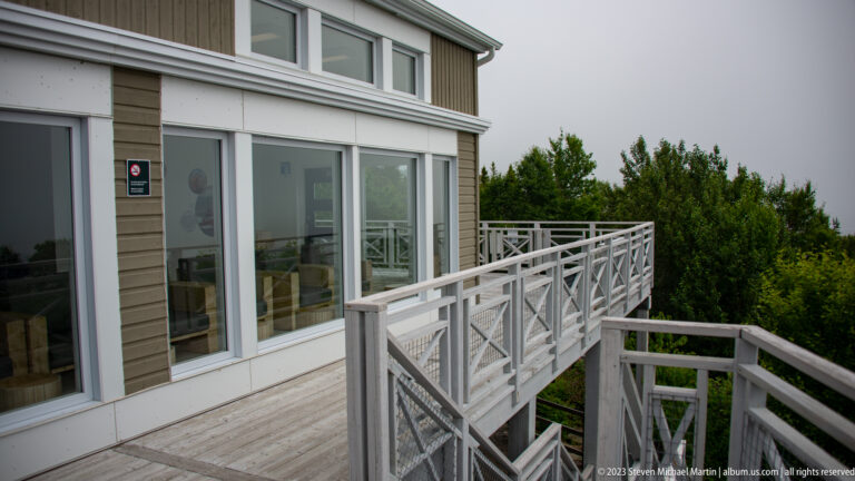 Light-sided museum building with large windows, wooden deck railings, and stairs above dense coastal vegetation.