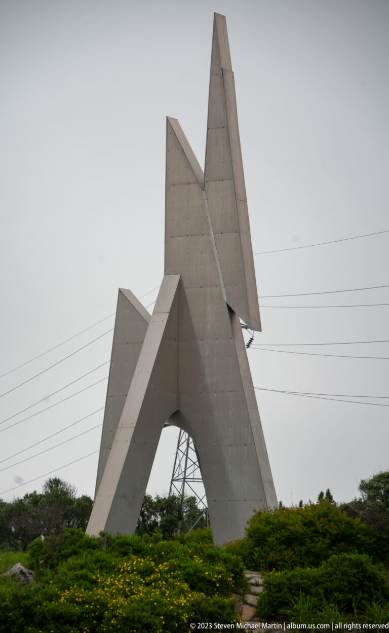 Large angular concrete sculpture with intersecting triangular forms rising above low shrubs beside utility lines.