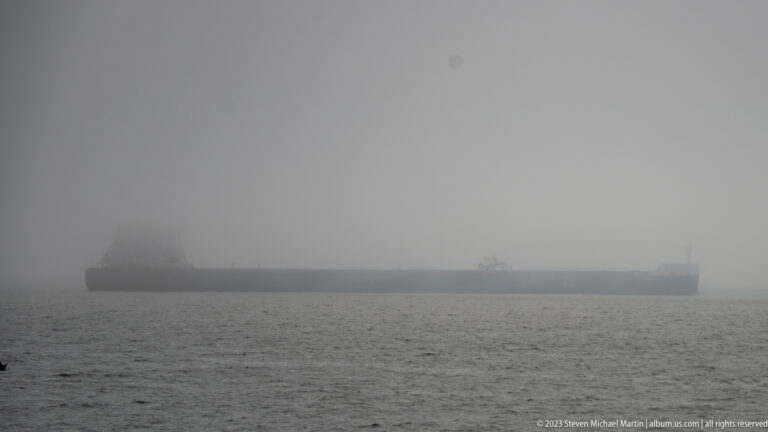Long dark cargo vessel barely visible through dense fog on gray coastal water.