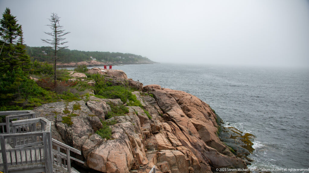 Rocky foggy shoreline with pink-gray bedrock, low shrubs, conifers, and a wooden stair at lower left.