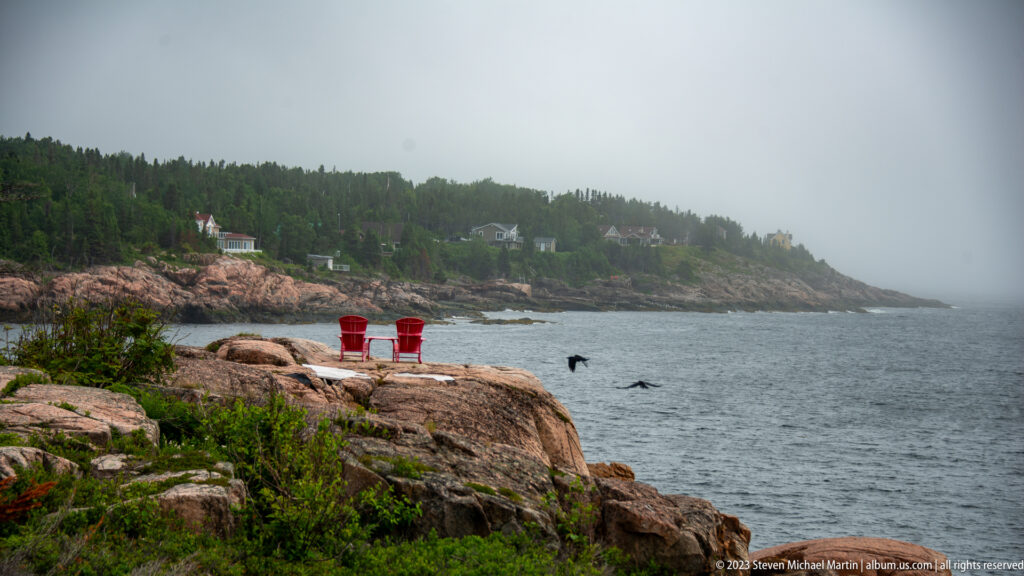 Two red Adirondack chairs on coastal rock overlooking a misty bay and forested shore, with a bird in flight.