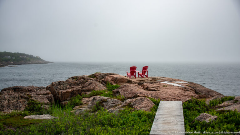 Wooden boardwalk through low shrubs leading to two red Adirondack chairs on foggy coastal rock.