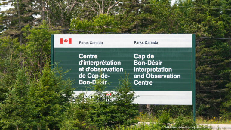 Green bilingual Parks Canada sign reading Cap-de-Bon-Désir Interpretation and Observation Centre among conifer trees.