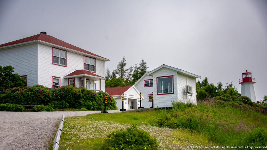 White red-trimmed station buildings on grass with the lighthouse rising behind them.