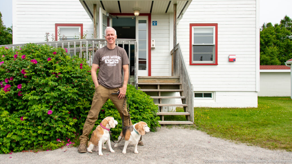 Man with two beagles standing by porch steps outside a white building with red window trim.