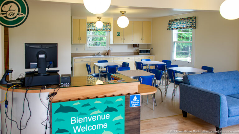 Bright room with welcome counter, tables, blue chairs, television, kitchenette, and curtained windows.