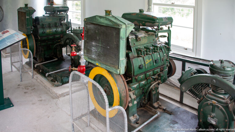 Two large green stationary engines with radiators, flywheels, pipes, and auxiliary machinery inside a white room.