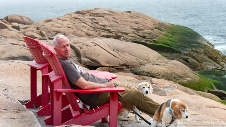 Man seated in a red Adirondack chair with two beagles on broad coastal rocks beside the sea.