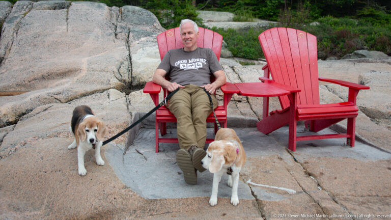 Man seated between two red Adirondack chairs with two beagles standing on the rock in front.