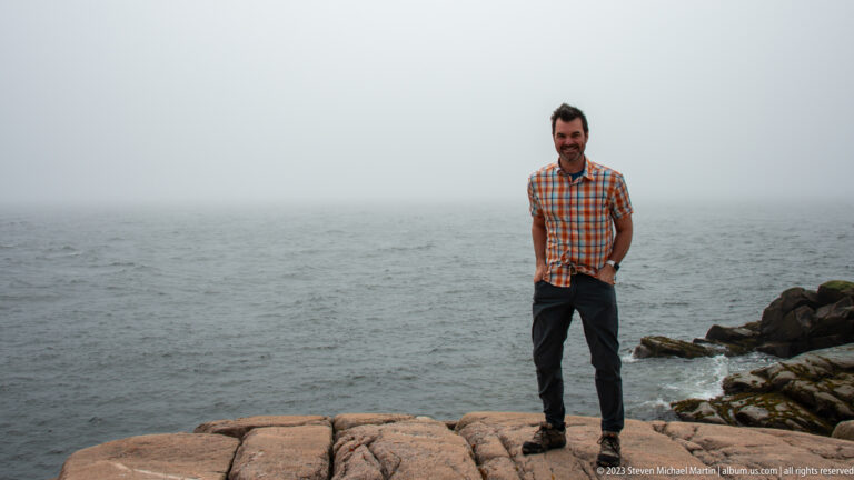 Man standing on pink-gray coastal rock beside a calm sea fading into fog.