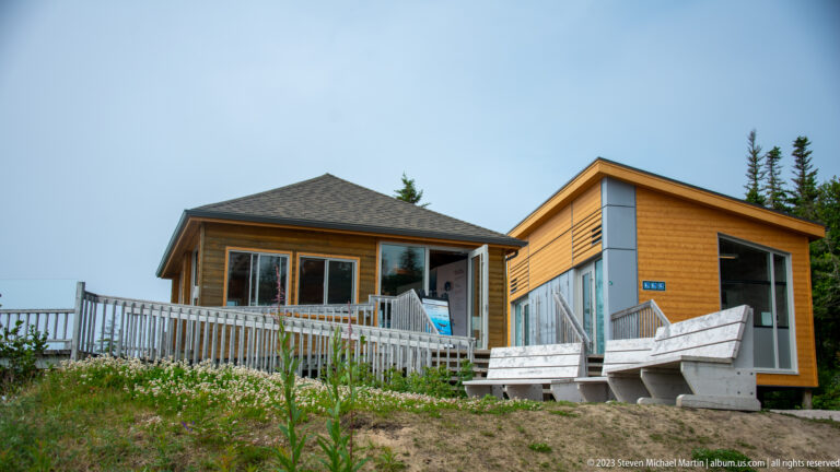 Small cedar-sided visitor building with large windows, accessible ramp, benches, and conifers.