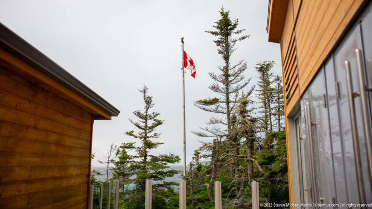 Canadian flag on a pole between cedar-sided buildings and narrow conifer trees.