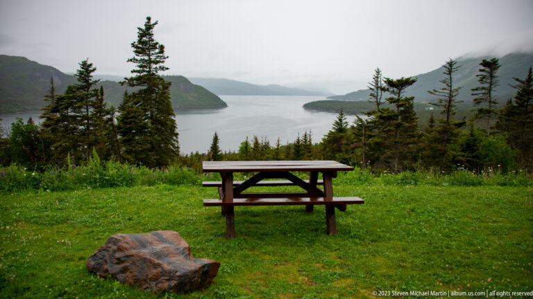 Wooden picnic table and reddish boulder overlooking a narrow mountain inlet framed by spruce trees.