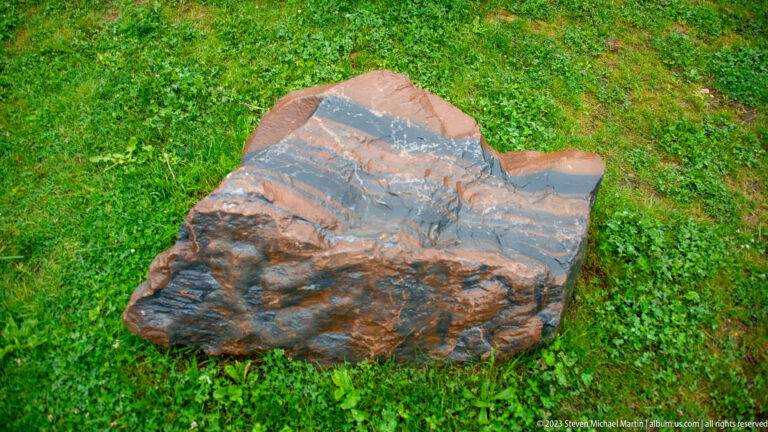 Large reddish-brown rock on grass with folded dark gray bands crossing its exposed surface.