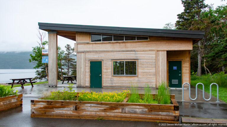 Small wood-sided park building with bike racks, flower beds, picnic tables, and water behind it.