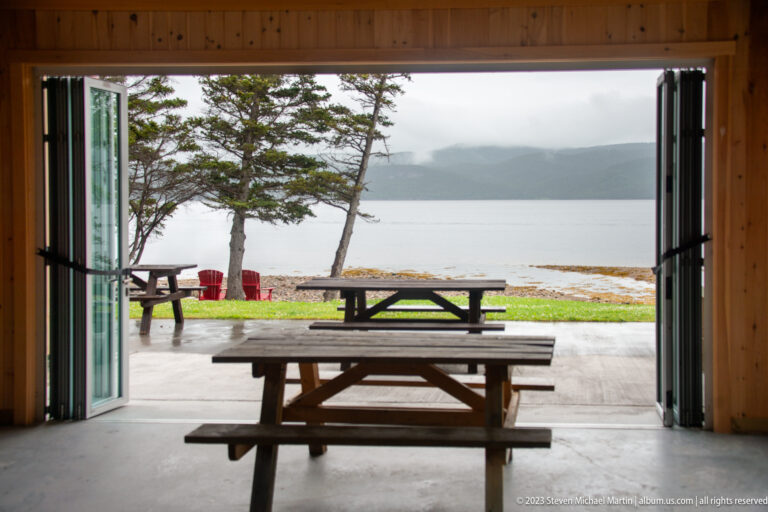 Picnic tables seen through open wooden doors toward red chairs, rocky shore, calm water, and misty hills.