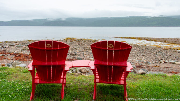 Two red Adirondack chairs on grass facing a rocky shore, calm water, and misty green hills.