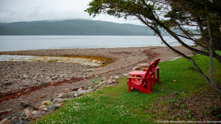 Two red Adirondack chairs beneath evergreen branches beside a curving rocky shore and calm water.