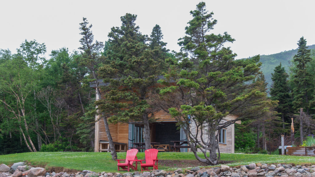 Wood-sided cabin among conifer trees with two red Adirondack chairs on a lawn beside a rocky edge.