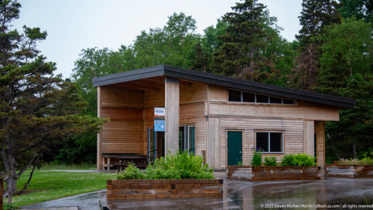 Modern wood-clad park building with a deep roof overhang, upper windows, planters, benches, and wet pavement.