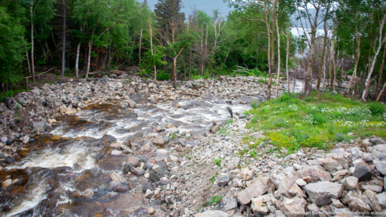 Whitewater stream flowing through a boulder-strewn channel with rocky banks and mixed forest on both sides.