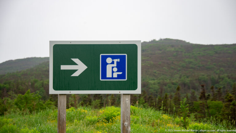 Green directional sign with a white arrow and blue binocular-viewpoint pictogram, set before low mountains and shrubs.