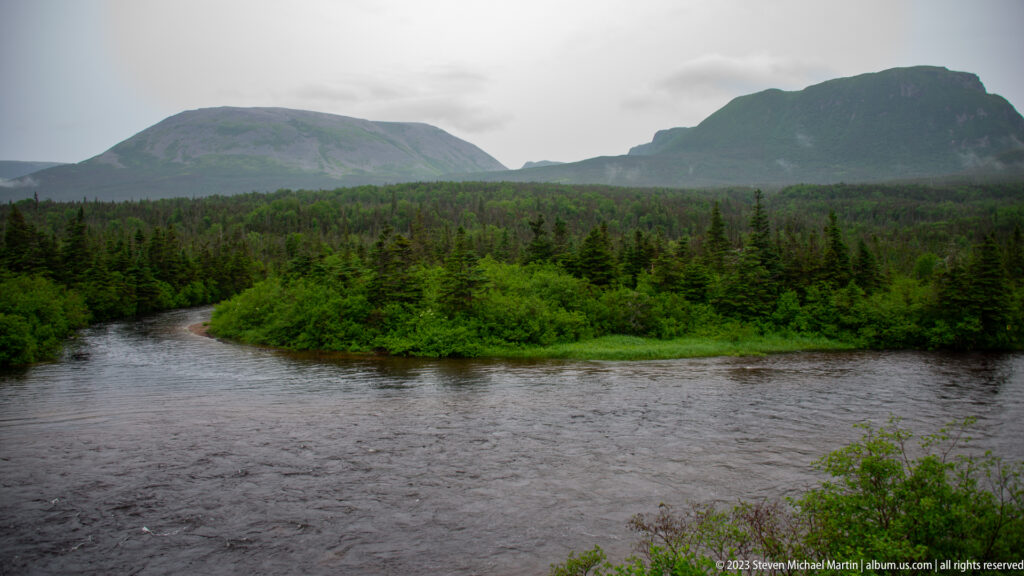 Broad river curving past forested land with two large mountains rising in the distance under gray skies.