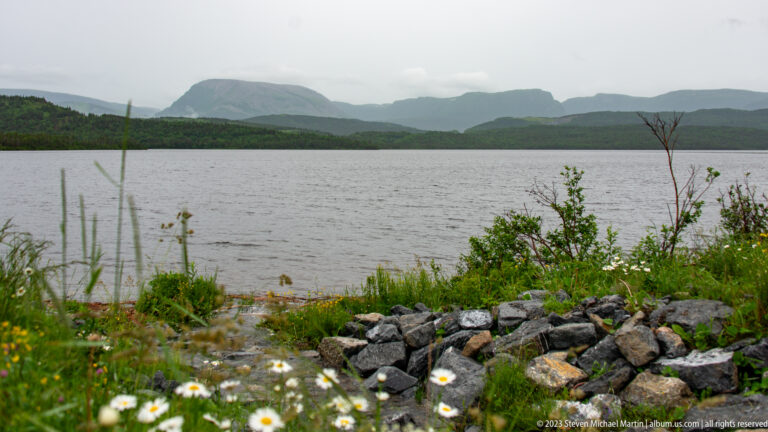 Calm inland water with a rocky, flower-dotted shoreline and layered mountains fading into low cloud.