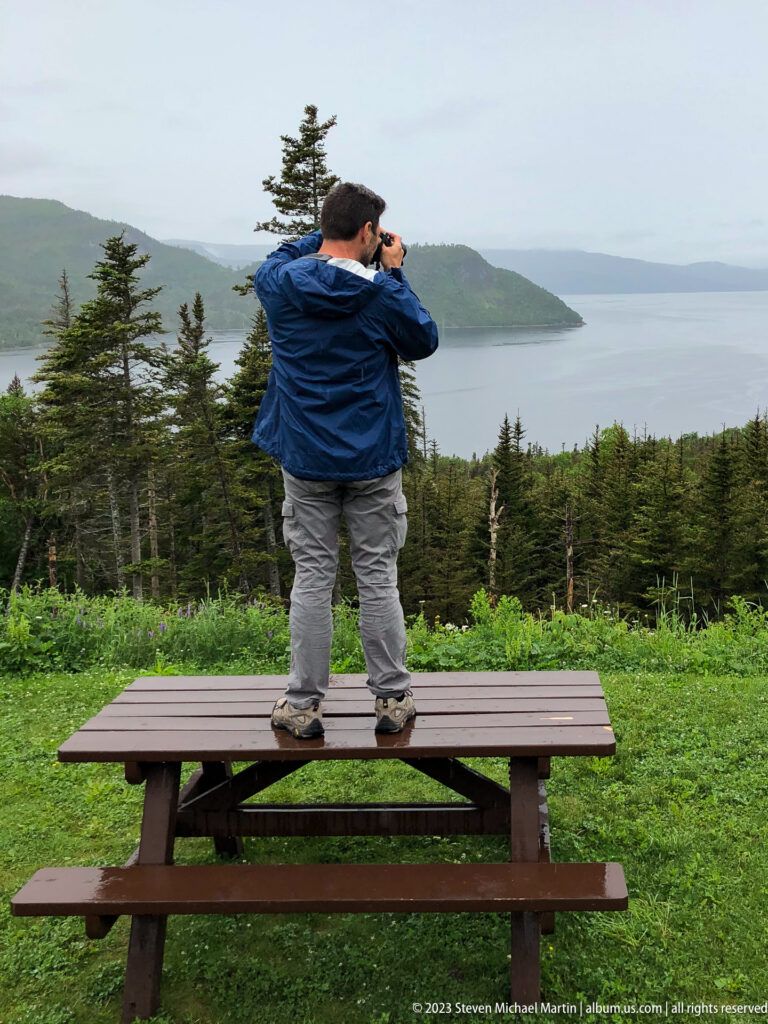 Person standing on a picnic table and looking through binoculars toward a broad inlet and distant mountains.