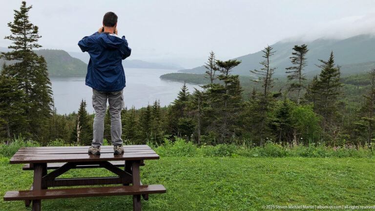 Visitor standing on a picnic table and using binoculars to look across a misty inlet framed by conifers and mountains.