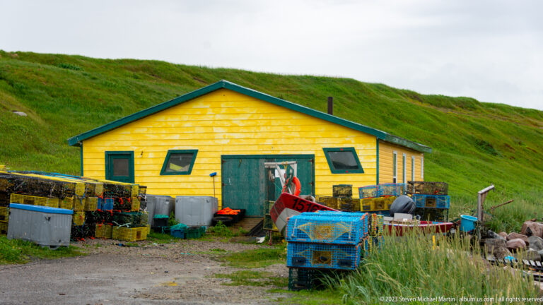 Yellow wood-sided work shed surrounded by fishing gear, crates, bins, and equipment at the base of a grassy slope.