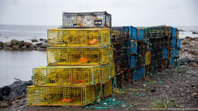 Stacks of yellow, blue, and black wire lobster traps with ropes and buoys on a rocky shoreline beside gray water.