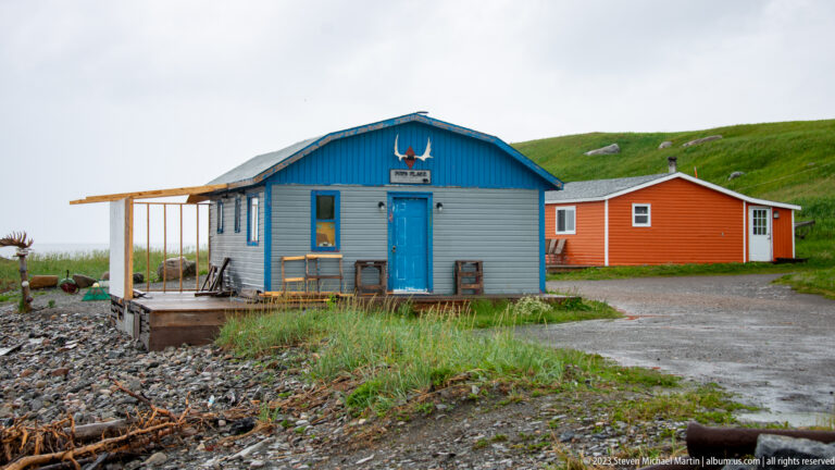 Small blue-and-white and orange-red coastal buildings on a rocky shore below a grassy hillside.